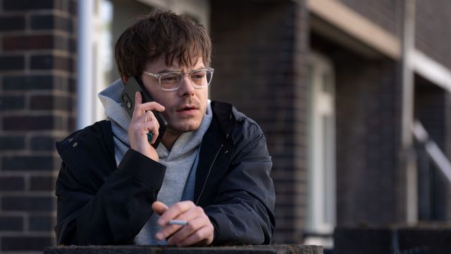 alt="Charlie Heaton as Jim Dycker speaking on a phone outside wearing glasses and a hoodie"