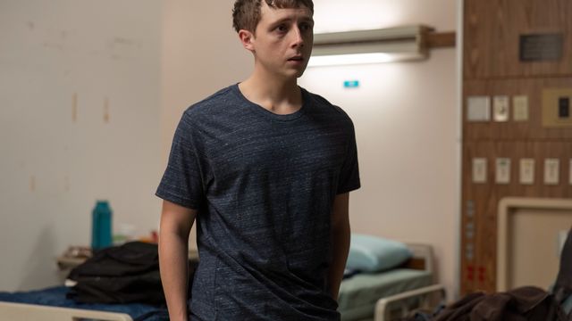 alt="Dennis Whitaker stands in a hospital room near a bed, looking anxious"