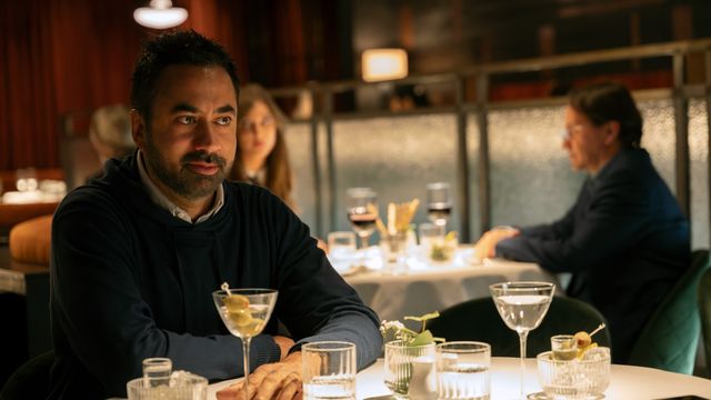 alt="Kal Penn as Jay Jonah Atterbury sits at a restaurant table with a martini, watching others in a dimly lit dining room."