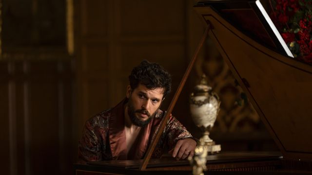 alt="Kit Harington as Sir Henry Muck leans over a piano in an ornate room, looking intense"