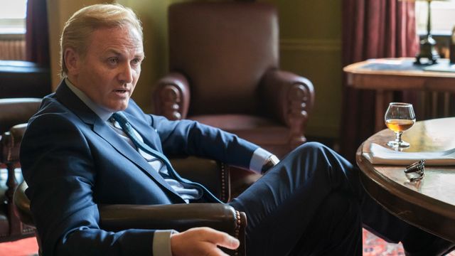 alt="Roger Barclay as Otto Mostyn in a leather chair, gesturing as he speaks to someone out of frame"